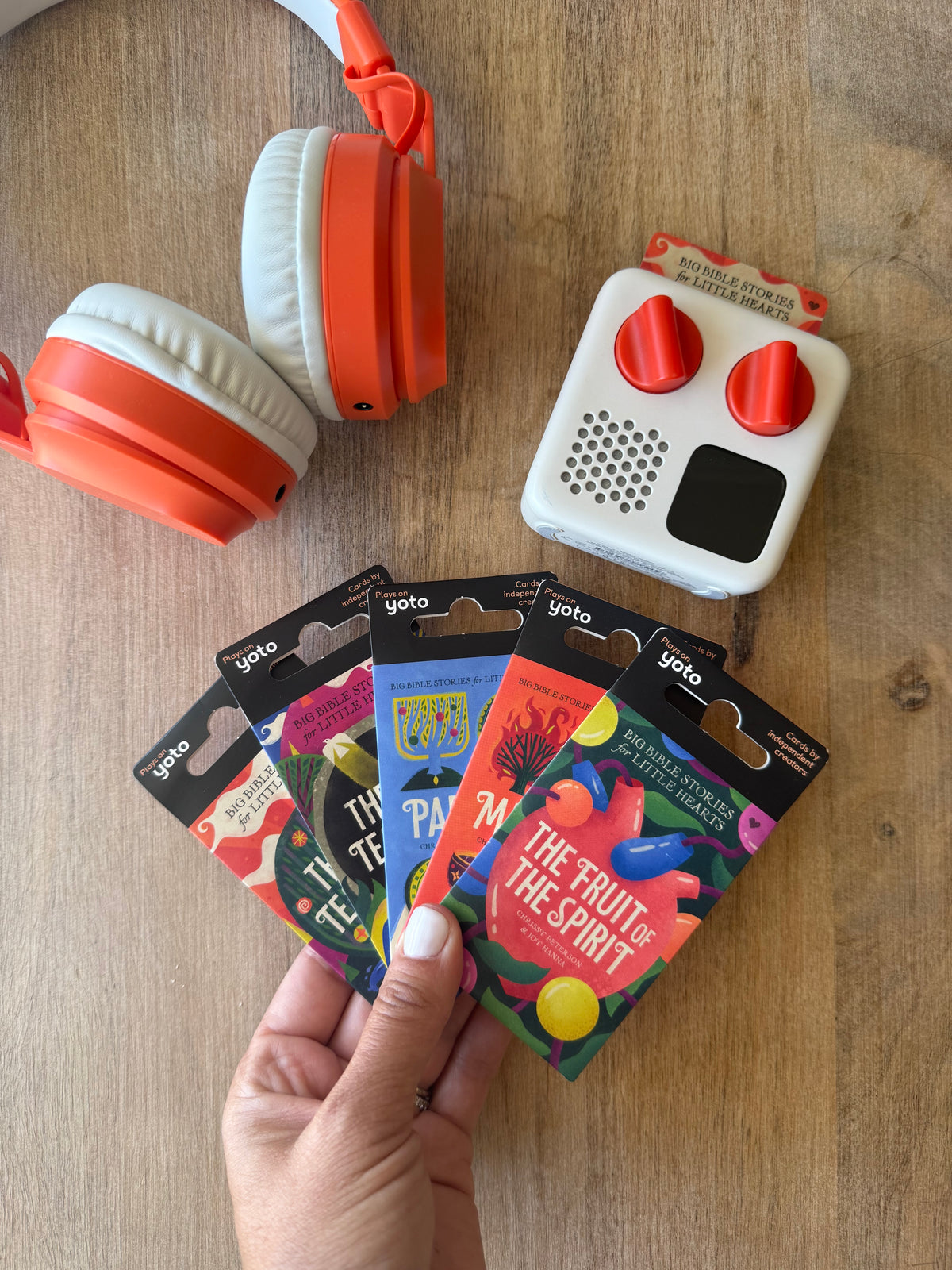 A hand holds a fan of colorful GQ magazine covers on a wooden tabletop, with orange and white over-ear headphones and a small white speaker nearby.