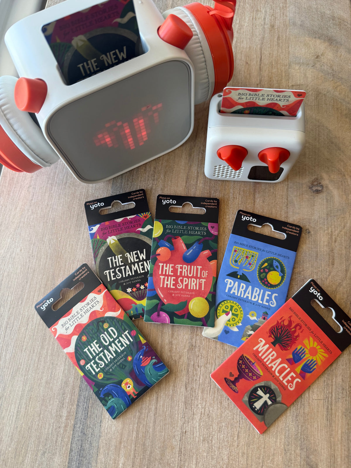 A set of Yoko brand religious card decks and a matching digital alarm clock arranged on a light wood tabletop. The card decks include 'The New Testament', 'The Fruit of the Spirit', 'Parables', and 'Miracles', while the clock features red knobs and a digital display.