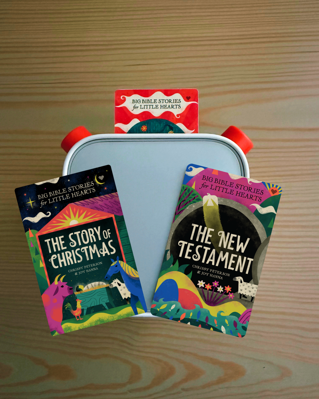 Three colorful children's Bible story books arranged on a white plastic tray with red handles, placed on a light wood surface. The books visible are 'The Story of Christmas', 'The New Testament', and 'Big Bible Stories for Little Hearts'.