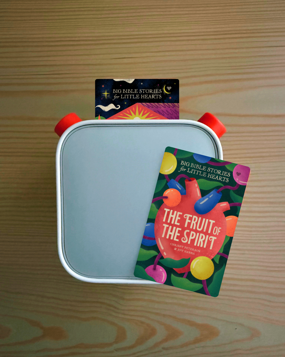 A square card game box with a light blue interior and red side handles sits on a wooden tabletop. A colorful card titled "The Fruit of the Spirit" is placed on top, with another card visible behind it.
