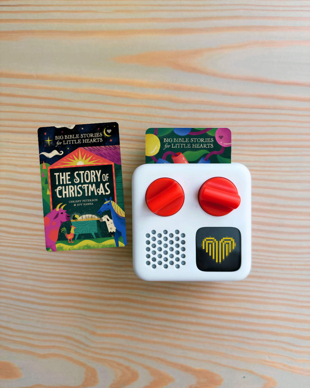 A white electronic card reader device with two red knobs and a speaker grille, shown next to its colorful 'The Story of Christmas' card on a light wood grain background.