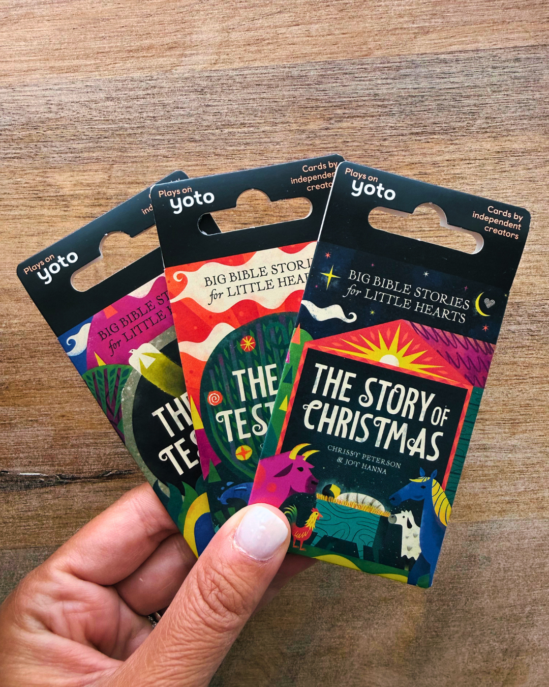 A hand holds three colorful Yoto Bible stories cards fanned out against a wooden tabletop background. The cards feature vibrant illustrations and titles including 'The Story of Christmas' and 'The Test'.