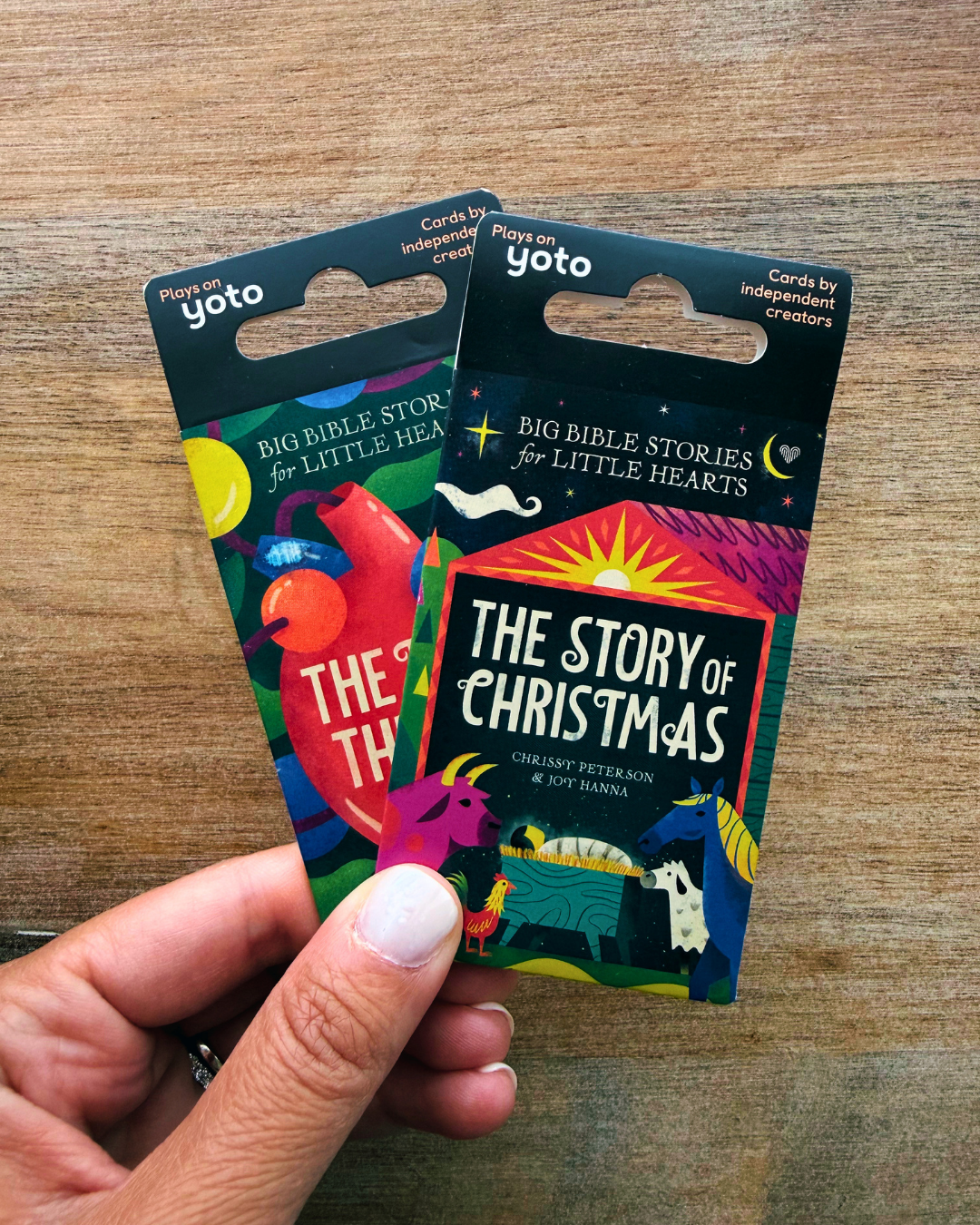 A hand holds two colorful Yoto religious cards, 'The Story of Christmas' and 'The Big Bible Story for Little Hearts', against a wooden tabletop background.